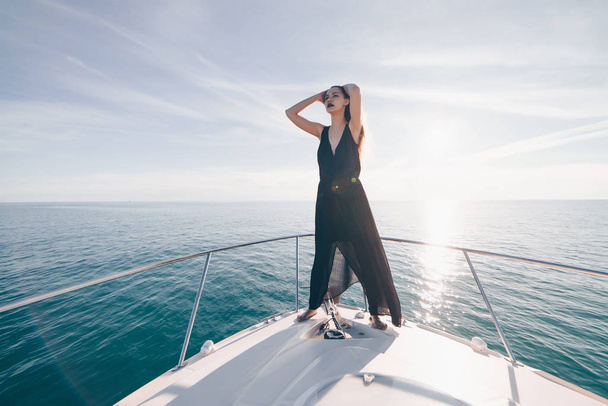 beautiful confident girl in a black summer dress enjoys sea voyage on her yacht in the rays of the sun - Foto, Imagem