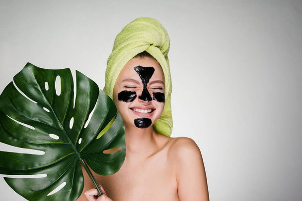 a laughing young girl cares for herself, on her face a black mask against acne, holds a green leaf - Photo, image