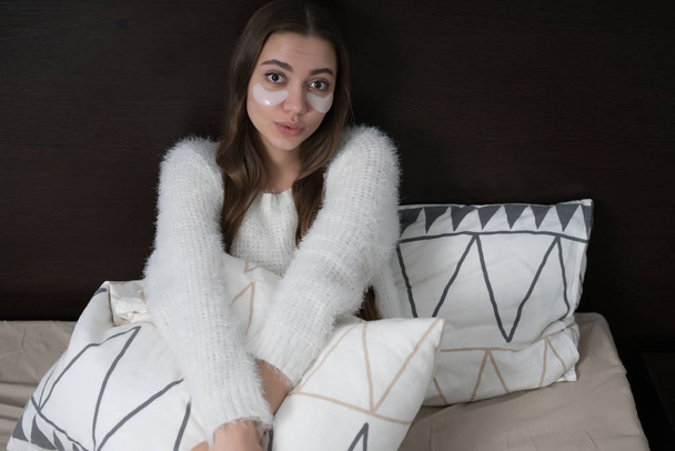 cute sleepy young girl in white pajamas sits in bed early in the morning, under the eyes white patches - Fotó, kép