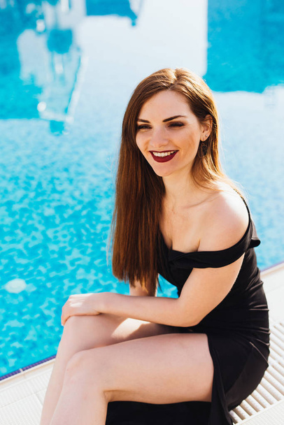 beautiful sexy young woman in black dress resting by the blue pool and laughing - Фото, изображение