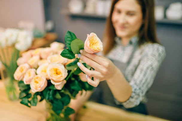 Θηλυκό ανθοπωλείο στην ποδιά βάζει ολόφρεσκα τριαντάφυλλα σε βάζο σε ανθοπωλείο, floral επιχειρηματική ιδέα - Φωτογραφία, εικόνα