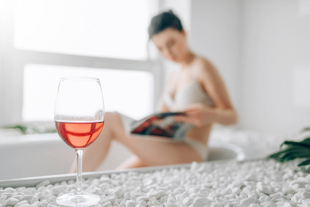 Young woman in white bathrobe sitting on the edge of the bath with foam. Bathroom interior with window and glass with red wine on background - Fotoğraf, Görsel