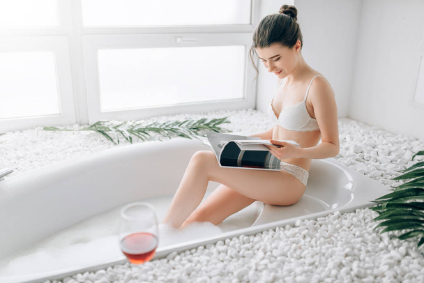 jovem mulher sentada no banho com copo de vinho tinto e revista de leitura. Interior do banheiro
 - Foto, Imagem