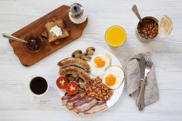 Bovenaanzicht, compleet Engels ontbijt met gebakken worstjes, eieren, bonen, spek en toast. Witte houten achtergrond. Van boven, overhead. Plat leggen.  - Foto, afbeelding