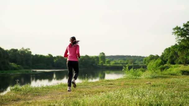 Dziewczyna w różowym kurtkę i czarne spodnie biegnie w pobliżu rzeki w słuchawkach przygotowania do maratonu - Materiał filmowy, wideo