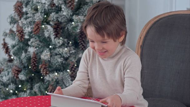 Smiling kid boy sitting on a chair and playing with tablet during Christmas time - Fotografie, Obrázek