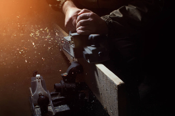 Foto de un trabajador procesando una madera con plano eléctrico con un viejo plano de acero de pie sobre vise con un fondo oscuro
. - Foto, imagen