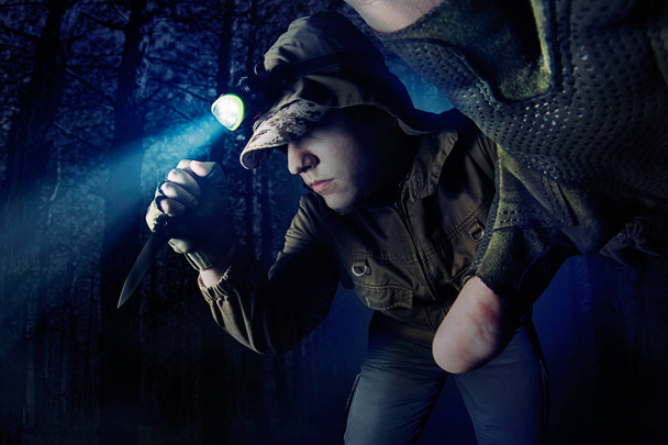 Foto de un hombre con chaqueta marrón táctica y guantes caminando en el bosque con linterna de cabeza y cuchillo táctico
. - Foto, imagen