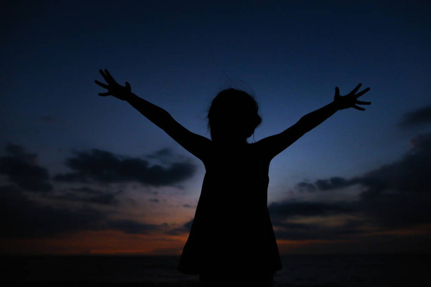 Silueta negra de niña con las manos extendidas en el fondo del cielo al atardecer
. - Foto, Imagen