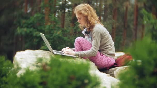 Kobieta z laptopa relaksuje się na skałach w pięknego, zielonego parku. Młoda kobieta wieloletnich w arboretum działa za laptopa. Technologia w plenerze - Materiał filmowy, wideo