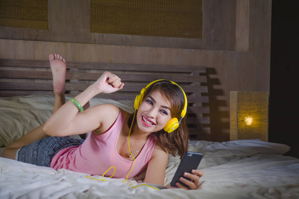 young attractive and happy girl in bed with yellow headphones listening to music on internet with mobile phone dancing and singing relaxed and cheerful in audio technology and lifestyle concept - Photo, image