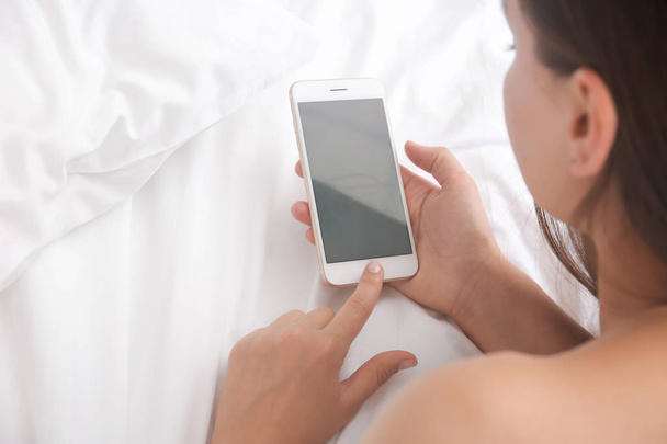Young woman holding mobile phone with blank screen in hand on bed - Fotó, kép