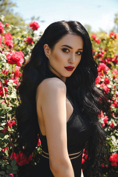 fashion outdoor photo of beautiful sexy woman with dark hair in elegant black dress posing in blossom garden with roses bushes - Photo, image
