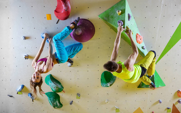 Rock climbers in bouldering gym climbing up overhanging wall. Young woman hanging upside down and smiling, male climber looking at her. - Photo, Image