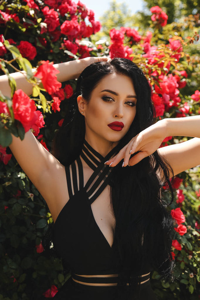 fashion outdoor photo of beautiful sexy woman with dark hair in elegant black dress posing in blossom garden with roses bushes - Фото, зображення
