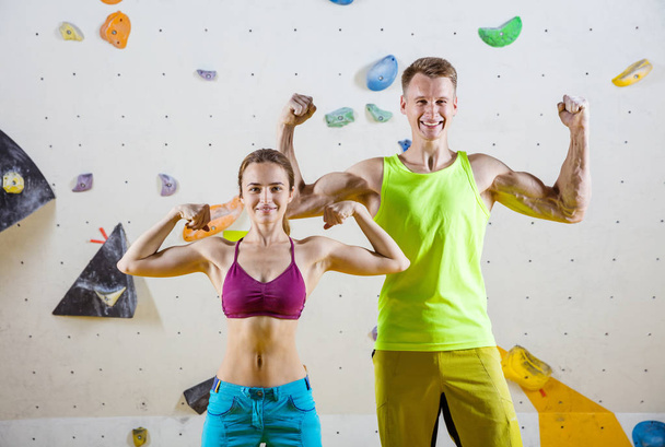 Young cheerful female and male rock climbers flexing biceps against bouldering wall in climbing gym  - Φωτογραφία, εικόνα
