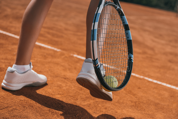 bijgesneden schot van vrouw oppakken van tennisbal met racket en been - Foto, afbeelding