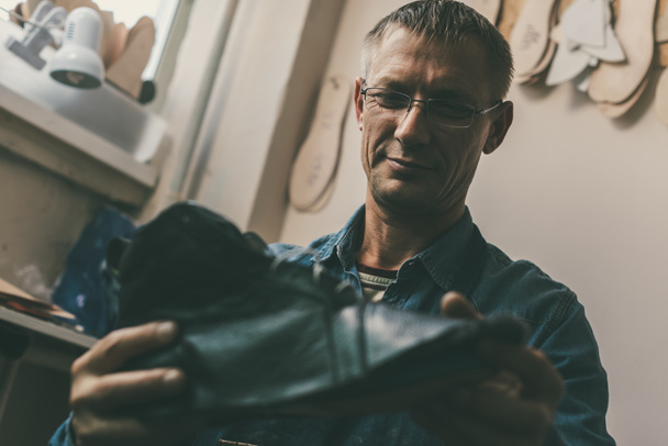selective focus of smiling mature cobbler in eyeglasses holding leather shoe in workshop - Photo, Image