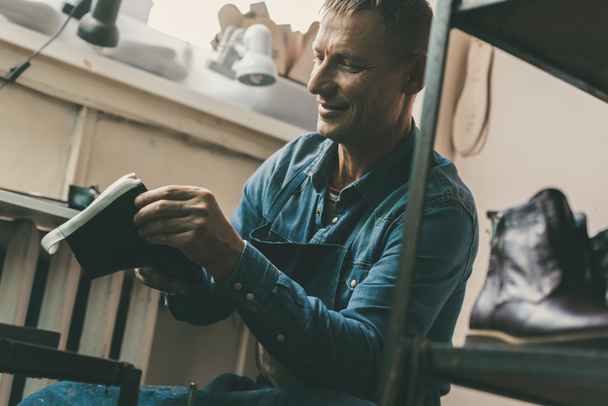 улыбающийся взрослый сапожник, работающий на пару обуви в мастерской
 - Фото, изображение