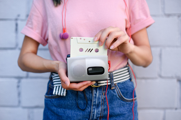 cropped shot of woman with earphones holding retro cassette player and audio cassette in hands against white brick wall - Photo, Image