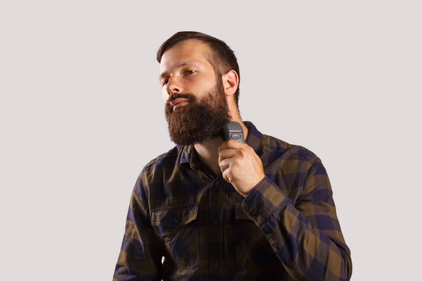 Így tökéletes a szakálla. Magabiztos fiatal szakállas férfi elektromos borotva borotválkozás, és látszó-on fényképezőgép, kockás ing, szürke háttérrel állva - Fotó, kép