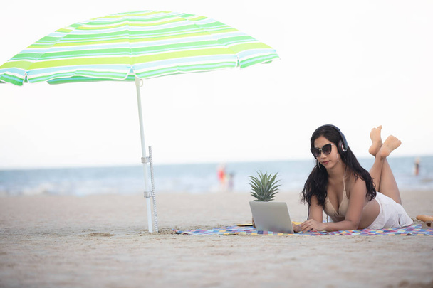 Γυναίκες φορώντας μπικίνι χρησιμοποιούν φορητό υπολογιστή για την εργασία και να ακούσετε μουσική στην παραλία το καλοκαίρι. - Φωτογραφία, εικόνα