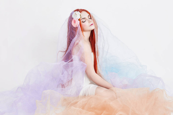 Redhead girl in light airy colored dress sits on the floor white background. Beautiful flowers in girl hair. Romantic woman with long hair and cloud dress. Girl dreaming bright makeup and perfect body - Foto, Imagen