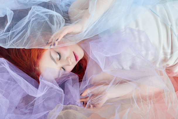 Redhead girl in a light air colored dress lies on the floor, a portrait close-up. Romantic woman with long hair and cloud dress. Girl dreams, bright makeup, clean skin, facial care - Photo, Image