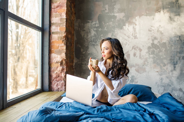 Petite brunette caucasian girl relaxing in bed with laptop in loft - Photo, Image