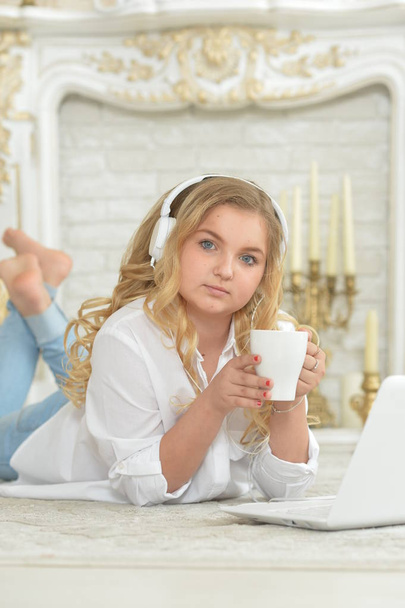 Curly blonde teenage girl in headphones lying on  floor with cup of tea and open laptop - Foto, imagen