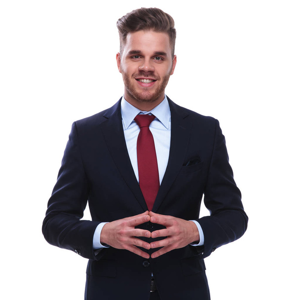 portrait of smiling young businessman standing on white background with fingers together. he is wearing a navy suit and a red tie - Фото, изображение