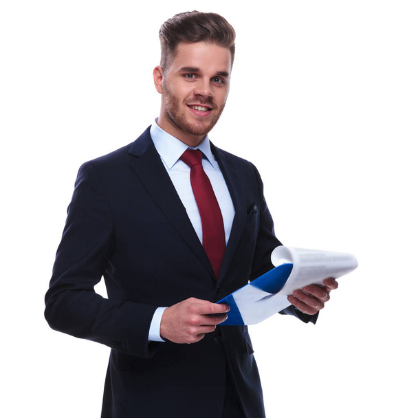 Πορτραίτο του νεαρός επιχειρηματίας, ελέγχοντας τα αρχεία του και χαμογελώντας ενώ στέκεται πάνω σε λευκό φόντο. Φοράει ένα Ναυτικό κοστούμι και κόκκινη γραβάτα - Φωτογραφία, εικόνα