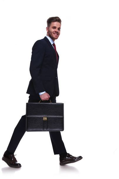 side view of businessman wearing a navy suit with suitcase in hand walking on white background, full body picture - Foto, Imagem