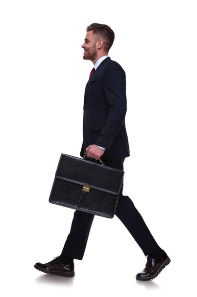 Zijaanzicht van de knappe jonge zakenman in Marine pak en met een koffer in de hand lopen als u wilt werken op witte achtergrond - Foto, afbeelding