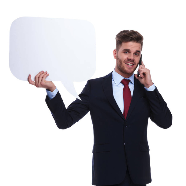 a telefon beszélő fiatal üzletember tart beszéd buborék állva fehér háttér, portré kép. Ő visel egy sötétkék öltönyt és piros nyakkendőt - Fotó, kép