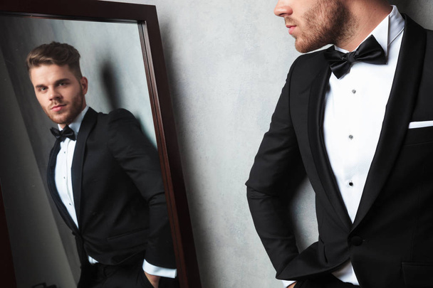 mirror reflection of a relaxed elegant young man wearing a black tuxedo while leaning against a white wall - Photo, Image