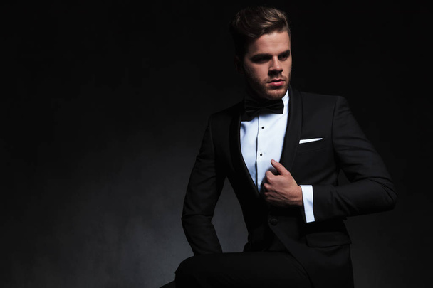 portrait of elegant young man in tuxedo looking to side while holding suit collar. He is sitting on a black background - Photo, Image