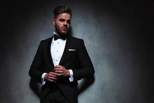 sexy man in black tuxedo holding hands together while looking to side and leaning against a dark grey wallpaper - Foto, immagini