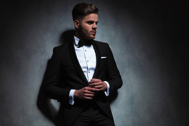 handsome unshaved man wearing black tuxedo looks to side while holding hands together and standing near a dark grey wall, portrait picture - Fotoğraf, Görsel