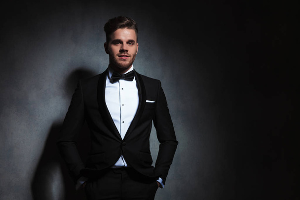 portrait of happy and relaxed young man in black tuxedo standing near a dark grey wallpaper with hands in pockets - Photo, Image