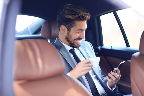 Businessman drinking coffee in car with phone in hand - Zdjęcie, obraz