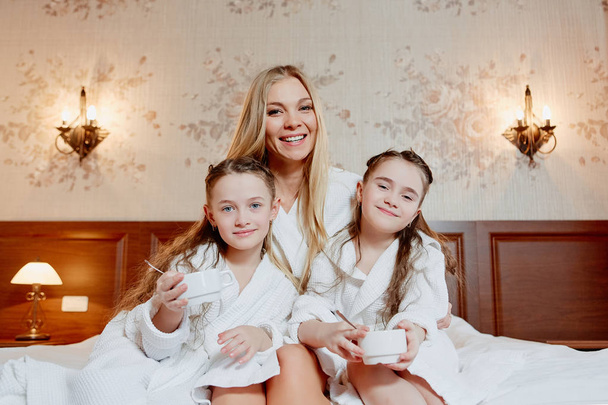 beautiful and happy mother and her two daughters, embrace and laugh. Sit and relax on a bed in the cozy room. Have breakfast. - Photo, image