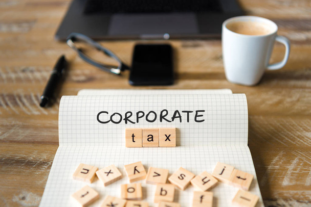Closeup on notebook over vintage desk surface, front focus on wooden blocks with letters making Corporate Tax text. Business concept image with office tools and coffee cup in background - Photo, Image