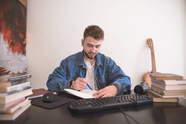 ハンサムな学生男コンピューターと本の山に座っていると、ノートの宿題で書きます。自宅ひげとスタイリッシュな学生。自己概念. - 写真・画像