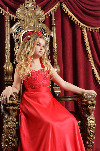 Blonde teenage girl in bright red dress sitting in vintage chair with red velvet curtains on background - Fotografie, Obrázek