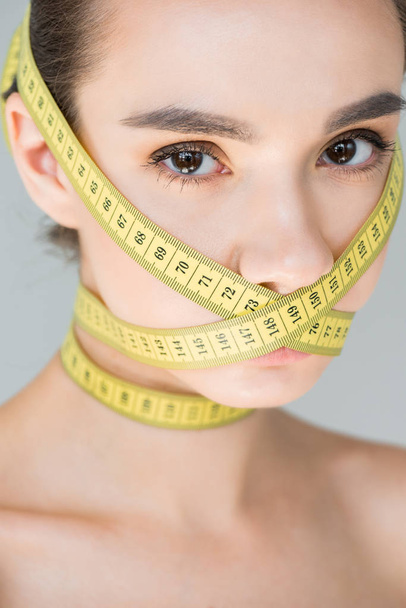 portrait of attractive young woman with closed mouth by measurement tape isolated on gray background  - Foto, Imagen