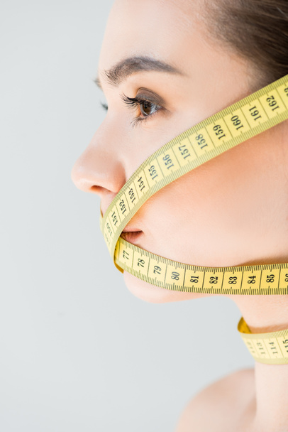 side view of attractive young woman with closed mouth by measurement tape isolated on gray background  - Foto, Imagen