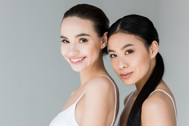 smiling asian and caucasian women looking at camera isolated on gray background  - Foto, imagen