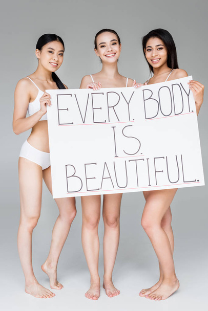 smiling multicultural women holding banner with lettering every body is beautiful isolated on gray background  - Фото, зображення