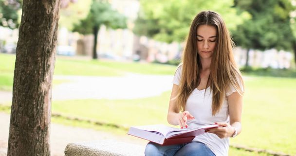Beautiful female college student reading a book on a bench in a park - Foto, imagen